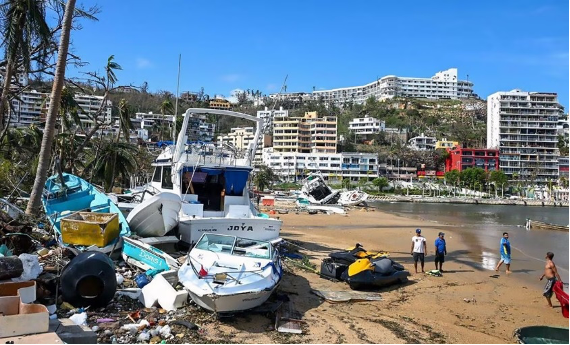 Un grupo de personas está de pie en una playa con barcos al fondo.