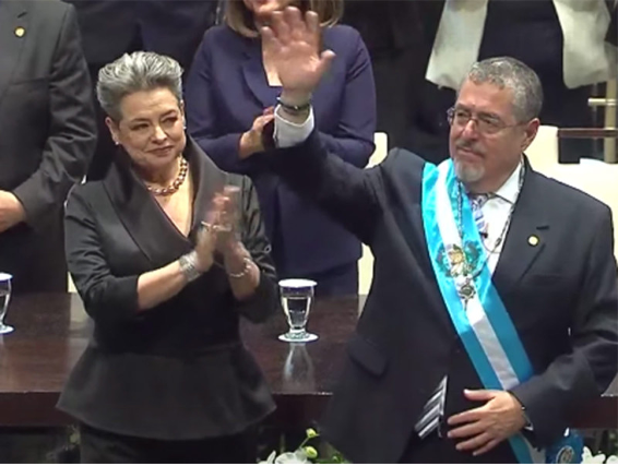 Un hombre de traje y una mujer de traje saludan.