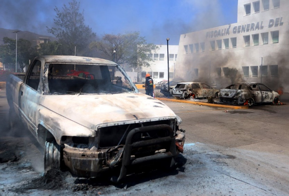 Bombero caminando cerca de vehículos quemados con humo de fondo.