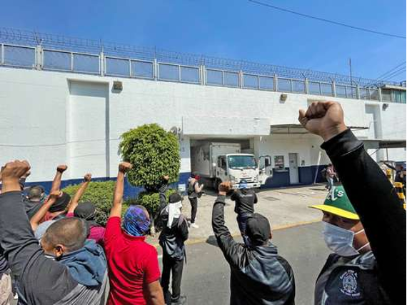 Individuos levantando el puño en gesto de solidaridad o protestando frente a un edificio con un autobús al fondo.