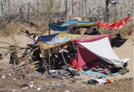 Refugio improvisado en una zona seca y escasa con varias telas colgadas para protección, rodeado de pertenencias personales dispersas.