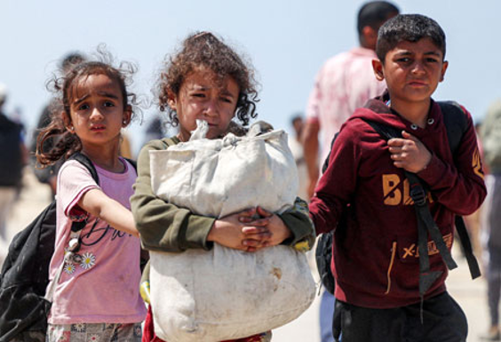 Tres niños vestidos con ropa informal caminan, uno de ellos cargando un gran saco y otro cargando una mochila, bajo un cielo despejado.