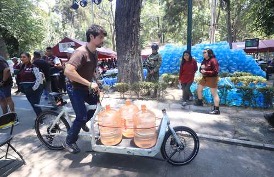 Un hombre transportando grandes botellas de agua de color naranja en un triciclo en un concurrido mercado al aire libre con gente y árboles.