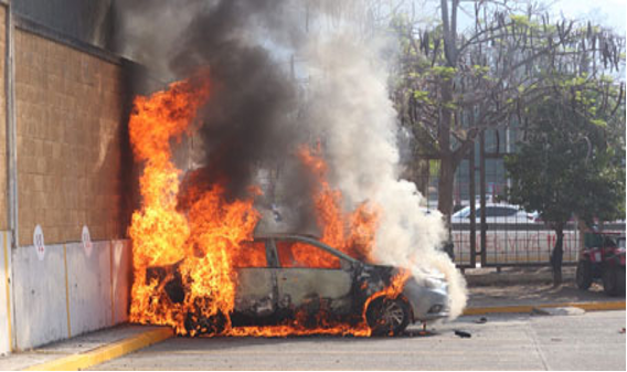 Coche envuelto en llamas junto a un edificio, emitiendo un espeso humo.