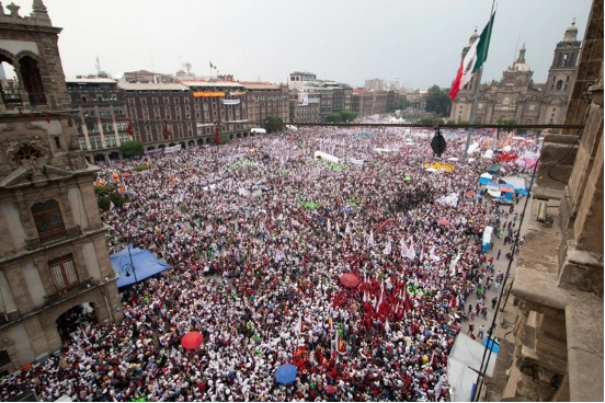 Una gran multitud llena una plaza de la ciudad frente a edificios históricos, con numerosos carteles y banderas de colores visibles.