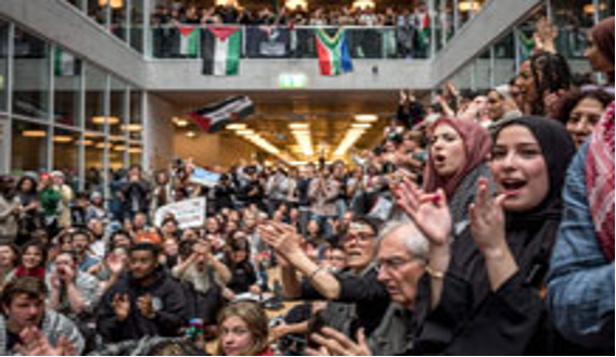 Diversa multitud de personas animando y aplaudiendo en un ambiente interior moderno con banderas y una escalera al fondo.