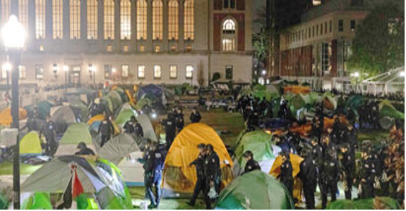 Numerosas tiendas de campaña y agentes de policía en un parque urbano muy iluminado por la noche, con un gran edificio al fondo.