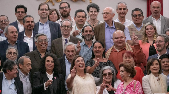 Un gran grupo de hombres y mujeres están juntos, sonriendo para una foto grupal en el interior. Varios individuos al frente levantan las manos y hacen gestos con los dedos.