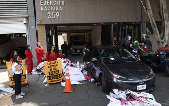 Un auto gris estacionado frente a un edificio con restos de papel esparcidos. Se ve a varias personas sosteniendo carteles y reuniéndose, algunas con chaquetas rojas. En el edificio se lee "EJERCITO NACIONAL 359.