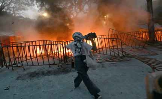 Una persona arroja un objeto hacia barricadas envueltas en fuego con nubes de humo, en medio de un fondo de árboles y una iluminación tenue.