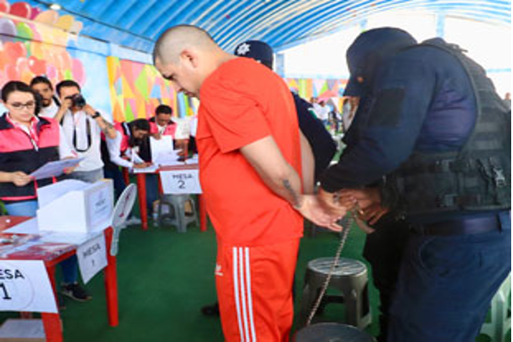 Un hombre esposado y vestido con un mono naranja es acompañado por un agente de policía en un mostrador de registro, con observadores al fondo.