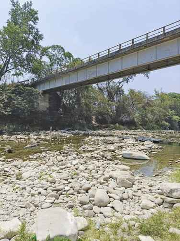 Un puente de metal se extiende sobre un lecho rocoso de un río con agua escasa. Altos árboles se alinean a las orillas del río bajo un cielo despejado.