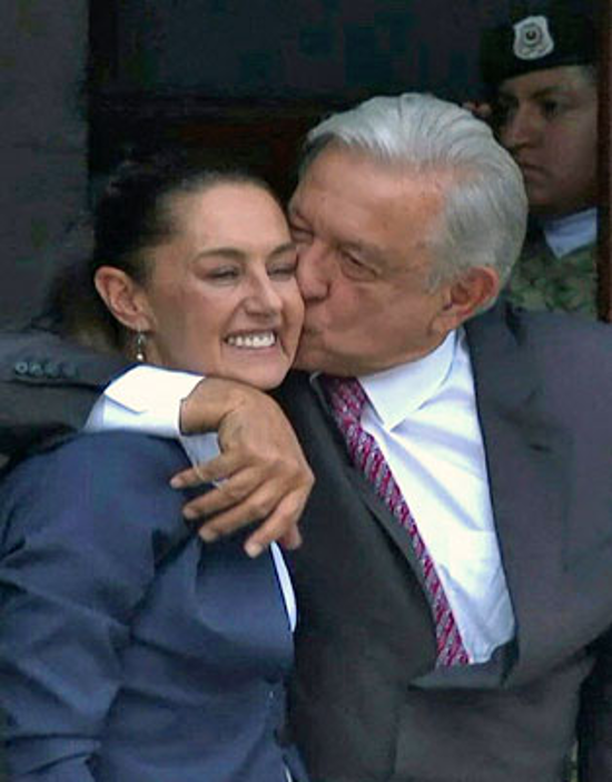 Un hombre de traje abraza y besa la mejilla de una mujer sonriente vestida de negocios. Al fondo se ve un guardia uniformado.