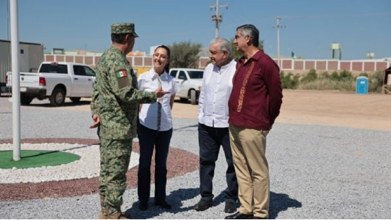 Un grupo de cuatro personas, incluida una persona con uniforme militar, conversan al aire libre sobre una superficie de grava con vehículos estacionados y edificios al fondo.