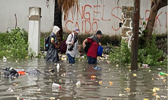 La gente camina a través del agua hasta las rodillas en una calle con escombros dispersos. Una pared al fondo tiene graffiti.