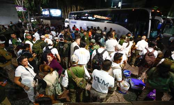 Un gran grupo de personas, entre ellos uniformados y civiles, se reúnen por la noche cerca de dos autobuses blancos. Algunas personas llevan mochilas y equipo, mientras que otras conversan.