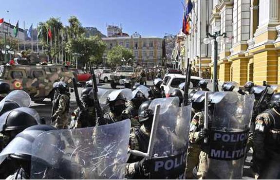 Una fila de policías antidisturbios con escudos se enfrenta a un grupo de vehículos militares en una calle de la ciudad con banderas y edificios al fondo.