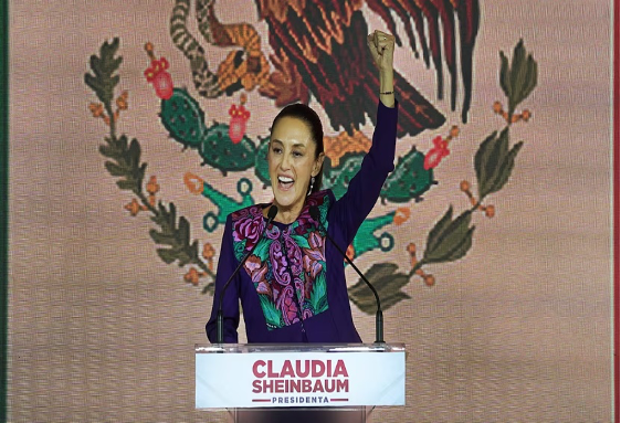 Una mujer se encuentra detrás de un podio con el cartel "Claudia Sheinbaum Presidenta", levanta el puño y habla, con el sello de México de fondo.