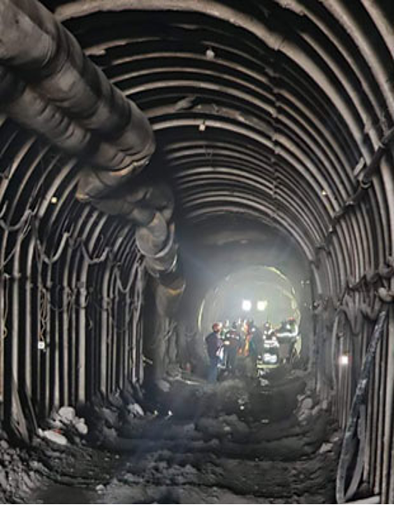 Los trabajadores se encuentran dentro de un túnel parcialmente construido, que cuenta con arcos de acero reforzado y está iluminado por luces brillantes en la distancia. Escombros y materiales de construcción están esparcidos por el suelo.