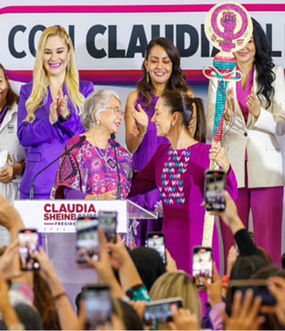 Un grupo de mujeres, algunas con teléfonos, rodean un podio donde dos mujeres hablan por micrófonos. Un cartel arriba dice "Claudia Sheinbaum". Una mujer sostiene un objeto ornamentado.