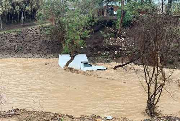 Un camión blanco está parcialmente sumergido en aguas fangosas, rodeado por un paisaje con árboles y colinas.