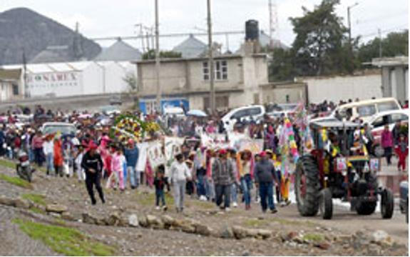 Un nutrido grupo de personas ataviadas con vestimentas coloridas participan en un desfile, caminando por una carretera con edificios y montañas al fondo. También se ven algunos vehículos, incluido un tractor.