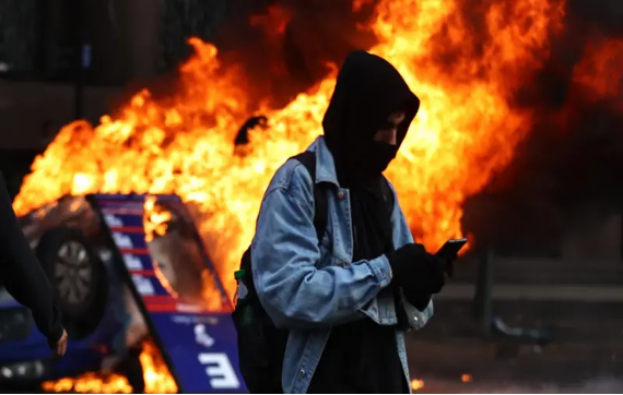 Una persona con una chaqueta con capucha y un cubrebocas utiliza un teléfono inteligente mientras un coche arde al fondo, envuelto en llamas.