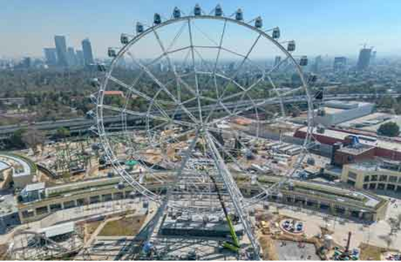 Vista aérea de una gran noria en un parque de diversiones con el horizonte de la ciudad al fondo. En los alrededores se pueden ver obras de construcción y otras atracciones.