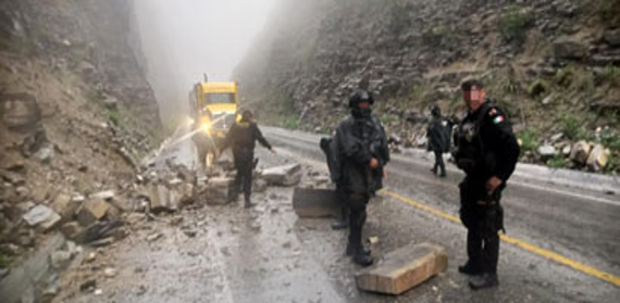 Los equipos de rescate y los vehículos limpian un deslizamiento de tierra en una carretera de montaña con niebla, con rocas y escombros esparcidos por el pavimento mojado.