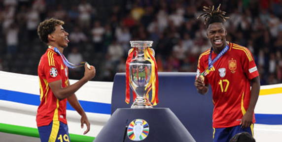 Dos jugadores de fútbol con uniformes rojos y amarillos celebran de pie junto a un gran trofeo en un podio, con medallas alrededor del cuello.