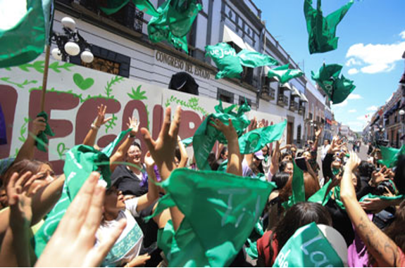 Una multitud de personas se reúne en una calle de la ciudad ondeando banderas y pancartas verdes en un evento al aire libre bajo un cielo soleado.