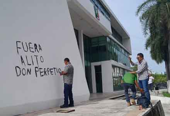 La gente destroza un edificio blanco, con dos hombres pintando con aerosol "FUERA ALITO DON PERPETUO" en la pared, mientras otro hombre toma una foto del graffiti.