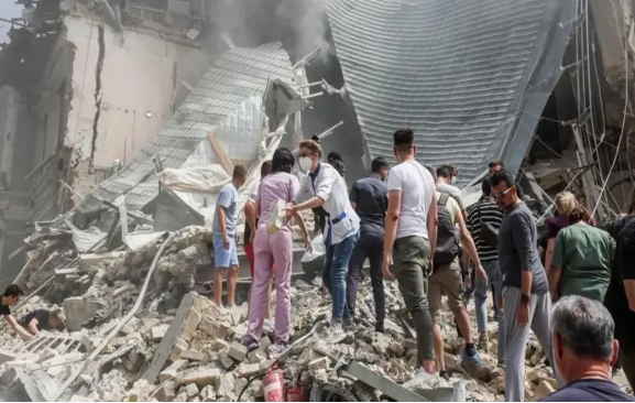 La gente se encuentra entre los escombros y los escombros de un edificio derrumbado, con humo elevándose al fondo. Algunas personas están ayudando en los esfuerzos de búsqueda y rescate.