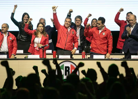 Un grupo de personas, en su mayoría vestidas con chaquetas rojas, están de pie en un escenario, algunas levantando los puños. En primer plano hay una multitud vitoreando con los puños en alto. En la mesa frente a ellos se ve el logotipo del "PRI".