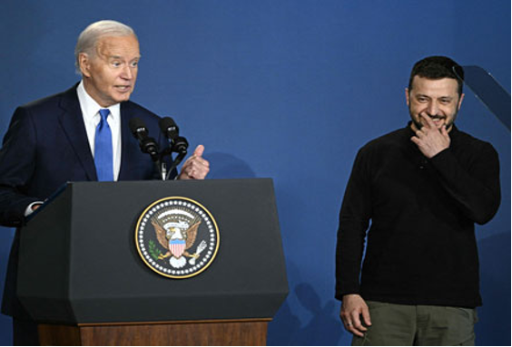 Un hombre de traje habla en un podio con el sello presidencial de Estados Unidos, mientras otro hombre vestido con ropa informal está de pie a su lado, sonriendo con la mano en la barbilla.