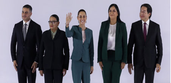 Cinco personas se encuentran en fila sobre un fondo liso. La persona en el medio sonríe y saluda. Los individuos visten vestimenta formal de negocios.