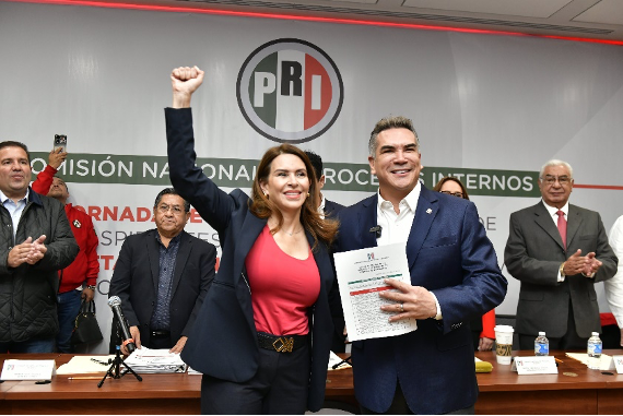 Dos personas juntas frente a una pancarta del PRI. Una persona levanta el puño en el aire, mientras la otra sostiene un documento. Otras personas se quedan al fondo, aplaudiendo y sonriendo.