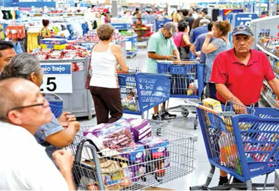 Un pasillo de supermercado abarrotado con numerosas personas haciendo cola en las cajas, empujando carritos de compras llenos de diversos productos. Al fondo se ven estantes con productos y carteles promocionales.