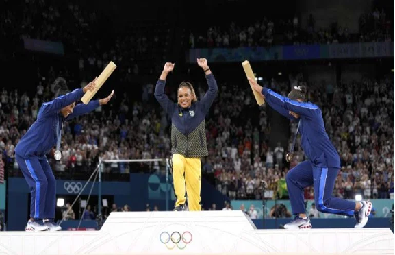 El atleta se para en el podio superior levantando los puños en señal de victoria, flanqueado por otros dos atletas a cada lado en los podios del segundo y tercer lugar, inclinándose con los brazos levantados.