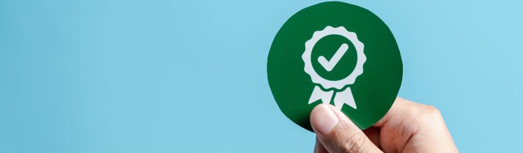 Mano sosteniendo un círculo verde con una marca de verificación blanca dentro de un símbolo de roseta blanca, que representa la formación continua y la certificación, sobre un fondo azul.