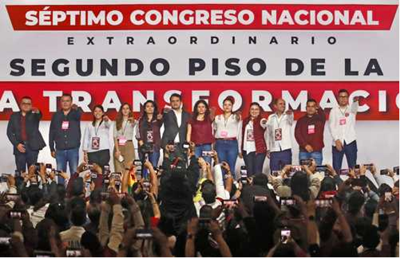 Un grupo de personas se encuentra en el escenario para una foto en el evento "Séptimo Congreso Nacional Extraordinario", con miembros de la audiencia fotografiándolos desde abajo.