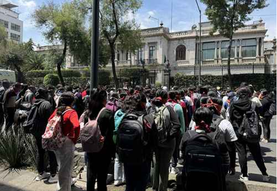 Un gran grupo de personas con mochilas y mascarillas se reúnen al aire libre cerca de un edificio histórico, bajo un cielo parcialmente nublado. Se ven árboles y otros edificios al fondo.