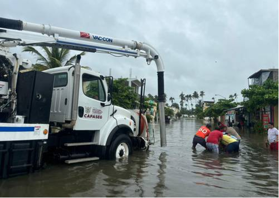 Un gran camión trabaja para bombear agua de una calle inundada, mientras tres personas con chalecos de seguridad ayudan en las aguas inundadas, rodeados de edificios y palmeras bajo un cielo nublado.