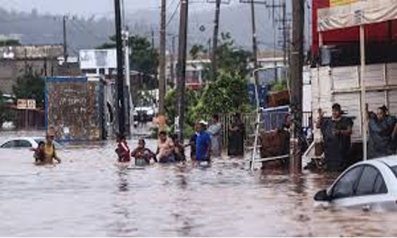 Gente caminando por una calle inundada con coches sumergidos en el agua y edificios al fondo.