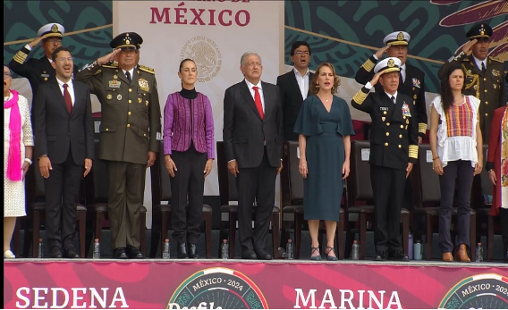 Un grupo de personas, entre ellas militares y civiles, se encuentran de pie sobre un escenario con un telón de fondo en el que se lee "México". Algunos saludan.