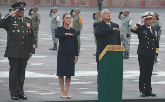 Oficiales saludando durante una ceremonia. Cuatro personas forman una fila: un hombre con uniforme militar, una mujer con traje, un hombre detrás de un podio y otro hombre con uniforme naval. Personal militar de pie al fondo.