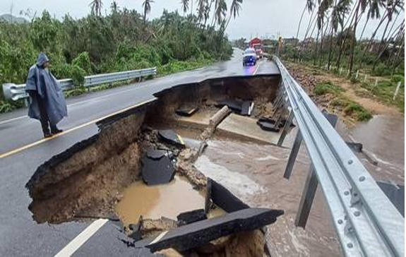 Una gran sección de la carretera se ha derrumbado y ha quedado cubierta de barro y agua. Cerca de allí hay una persona con impermeable y a lo lejos hay vehículos de emergencia estacionados.