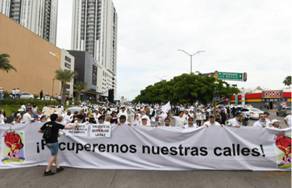 Un gran grupo de personas marcha por una calle de la ciudad con una pancarta que dice "¡Recuperemos nuestras calles!". Al fondo se ven algunos edificios y árboles. Los participantes visten ropa blanca.