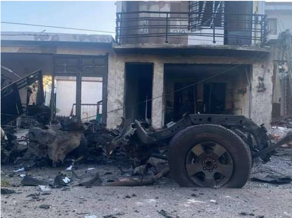 Restos carbonizados de un vehículo frente a un edificio dañado, con escombros esparcidos en el suelo.