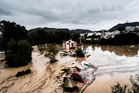 Paisaje inundado con coches parcialmente sumergidos y un edificio rodeado de agua fangosa. Al fondo se ven un cielo nublado y colinas.